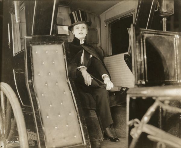 Marguerite Clark is seated in a Hansom cab in a publicity still for "The Amazons." She wears a man's tailcoat with white tie, cape, and top hat.
