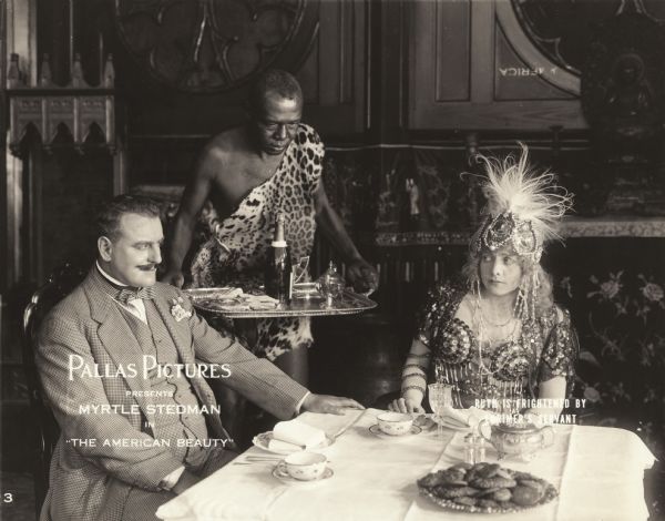 The dissolute Herbert Lorrimer (played by Howard Davies) leers at Ruth Cleave (Myrtle Steadman) as they are served champagne by an African servant wearing a leopard skin in a scene still for the silent drama "The American Beauty." Ruth wears a jewel and pearl encrusted dress and a gaudy headdress.