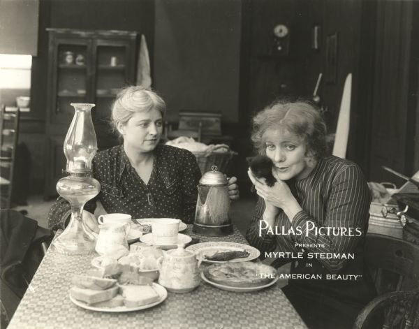 Mrs. Cleave (played by Adelaide Woods) and her adopted daughter Ruth (Myrtle Stedman) are seated for a meal in their humble home in a scene still for "The American Beauty." Ruth cuddles a black kitten. On the table are a pie, slices of bread, a battered coffee pot, and a kerosene lamp.