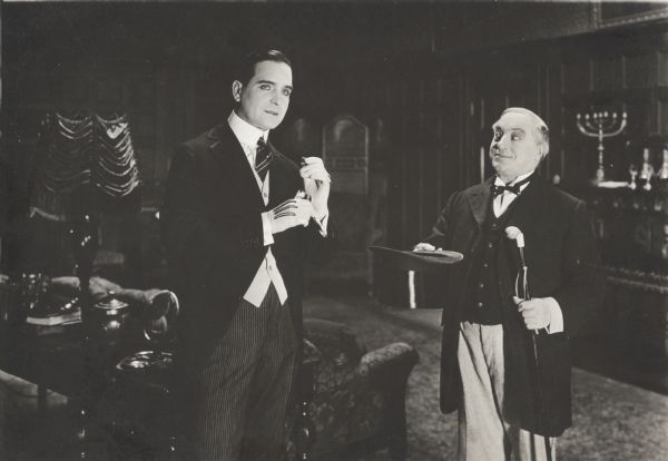 The silent film actor Earle Williams is offered a top hat and cane by a butler in a scene still. The film may have been "An American Live Wire" (Vitagraph 1918).