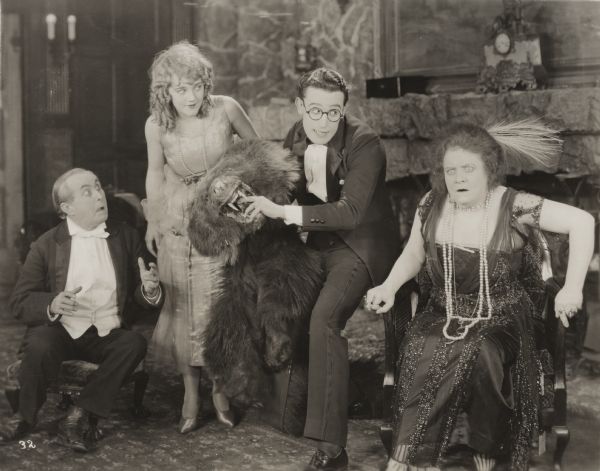 With the aid of a bear skin rug, Harold Lloyd tells an alarming hunting story in a scene still for "Among Those Present." From left to right the actors are James T. Kelley, Mildred Davis, Harold Lloyd, and Aggie Herring.