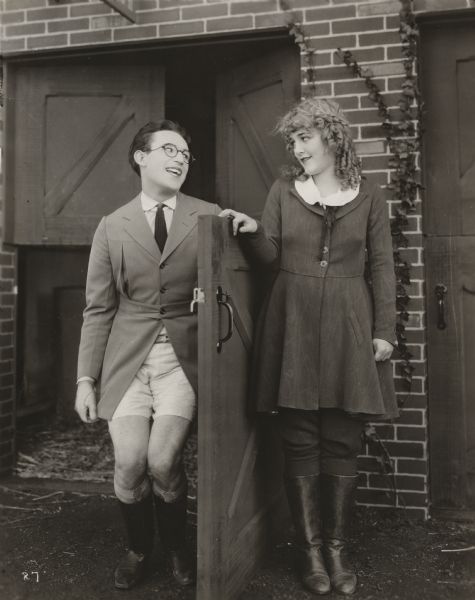 Harold Lloyd and Mildred Davis flirt at the stable door. Both wear riding clothes, but Lloyd has managed to lose his jodhpurs.