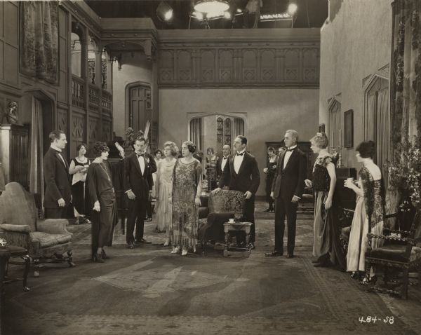 At least fifteen actors wearing formal evening wear pose in the drawing room of a mansion in this scene still for the silent film "Anna Ascends." In the first rank, from left to right the players are Robert Ellis, Alice Brady (with left arm raised), David Powell, Nita Naldi (in a complicated lace dress), and Charles Gerrard (with his hands on a chair back). A studio technician can be seen above the set tending the lights.