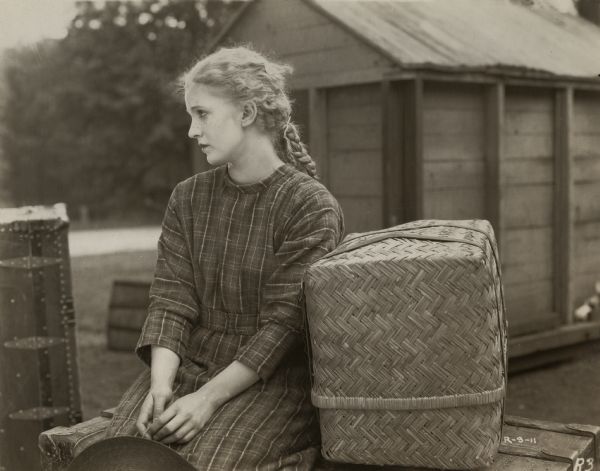Mary Miles Minter plays the orphan Anne Shirley in a scene still for "Anne of Green Gables." She is costumed as a young girl with pigtails and freckles and sits looking forlorn on a trunk with a basket beside her. The original caption written in pencil on the reverse of the print reads: "Anne has just come from the orphan asylum—but it looks like a case of the popular story 'Nobody knows and nobody seems to care.'"