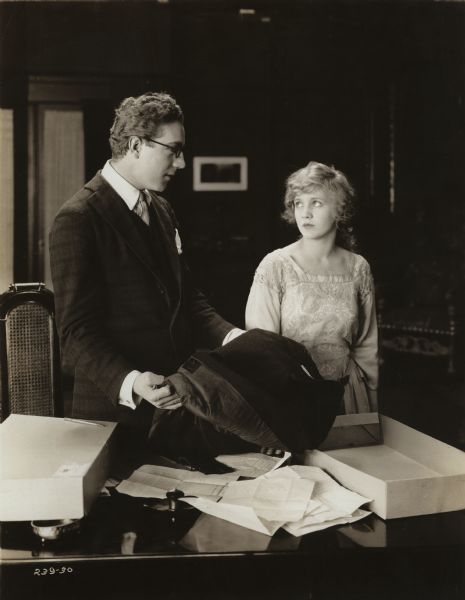 Willard Kaine Nottingham (played by George Fisher) removes a coat from a box and looks at Annie Johnson (Mary Miles Minter) in a scene still from the silent film "Annie for Spite" (Mutual 1917).