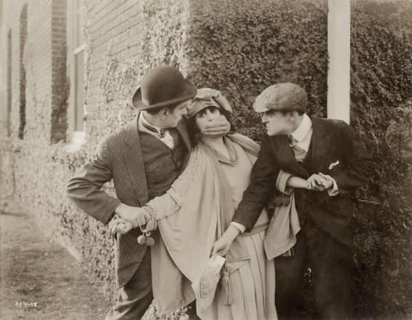 Ann Anderson (played by Margarita Fischer) is accosted by two ruffians in an outdoor scene still from the 1918 Mutual production "Ann's Finish." The man in the striped suit and derby holds a chloroform-soaked handkerchief on her mouth while the man in the cloth cap takes a letter from her pocket.