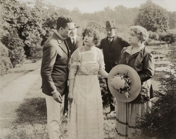 This outdoor scene still with the actors Robert Elliot, Dorothy Dalton, and Alice Gale in the foreground was made for the Thomas H. Ince production "L'Apache." The handwritten caption on the reverse of the print reads: "He would help her but why this mystery? Why was she silent?"