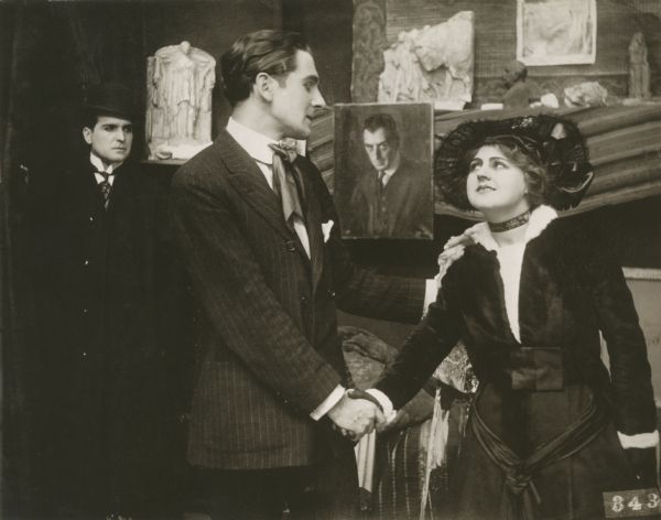 The actors Earle Williams, Rex Ingram, and Lillian Walker pose in a scene still from Vitagraph's "The Artist's Great Madonna." The scene is an artist's studio. Earle Williams is in the background among plaster casts while Rex Ingram, wearing the floppy tie of an artist, shakes hands with Lillian Walker wearing fashionable street clothes and a hat.