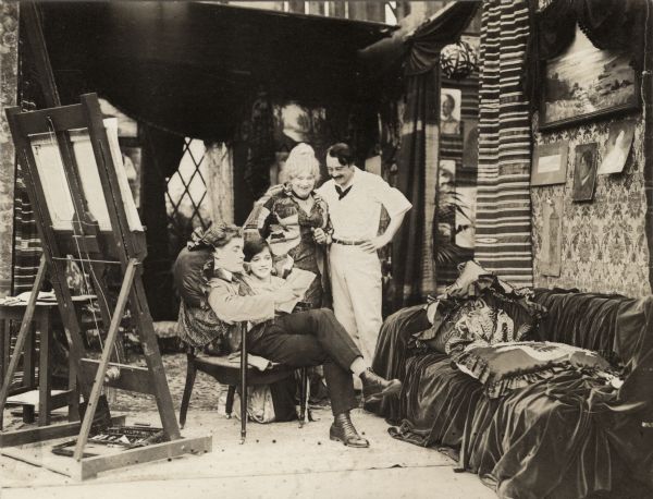 In this scene still for "The Artist's Wife," the young artist Adair (played by Elmer Clifton) is seated and holds a sketch or document in his hands. His model Jean (played by Miriam Cooper) kneels beside his chair while the actors Jennie Lee and Vester Pegg (probably in the role of George, Adair's friend) stand and look down benevolently at the young couple. They are in an artist's studio with a large easel and velvet covered posing couch. The room is decorated with paintings, sketches, and hangings of exotic fabrics.