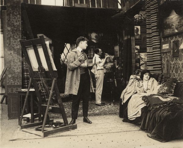 The young painter Adair (played by Elmer Clifton) stands by his easel holding his brush and palette. He is dressed in standard artist's garb: smock, floppy tie, and sash instead of a belt. In the background is his friend George (played by Vester Pegg). His model Jean (played by Miriam Cooper) wears an oriental robe and lies on a velvet couch with many cushions.