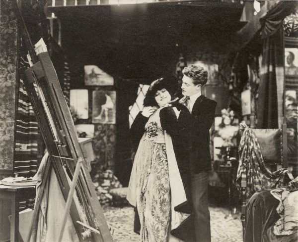 The young painter Adair (played by Elmer Clifton) helps a girl from high society (Lucille Young) with her wrap. They stand in his artist's studio.