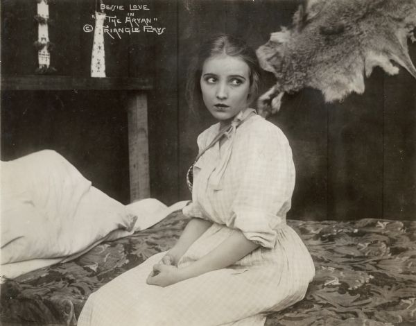In a scene still for the silent film "The Aryan," Bessie Love sits on a bed in a rustic room in her role as Mary Jane Garth, an innocent young girl crossing the wilderness with a wagon train.