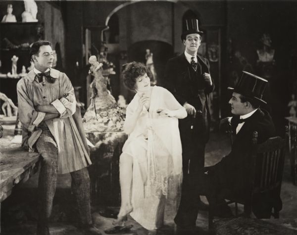 This scene still for "As a Man Thinks" is set in an artist's studio in Paris. On the left wearing a painting smock and floppy tie is the artist Benjamin De Lota (played by Warburton Gamble). Mimi Chardenet (played by Elaine Amazar) is his model. Seated wearing evening clothes and top hat is Frank Clayton (played by Henry Clive). The standing actor is unidentified.