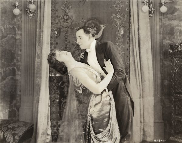 Elinor Clayton (played by Leah Baird) is being swept off her feet by the unscrupulous artist Benjamin De Lota (Warburton Gamble) in a scene still for the silent film "As a Man Thinks." Both are dressed in formal evening wear.