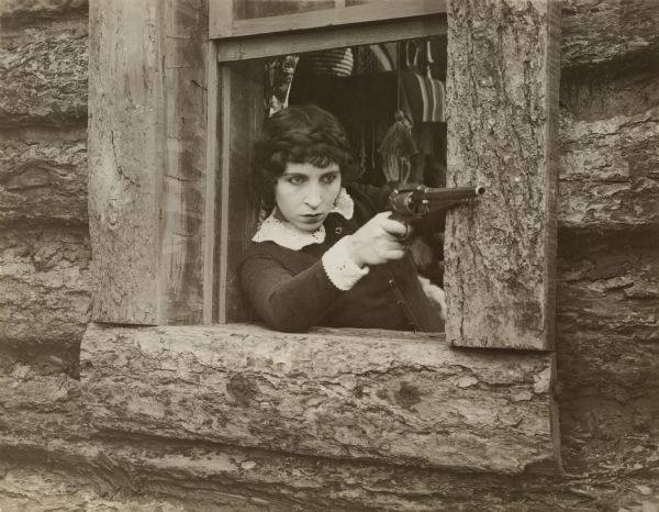 Edith Storey, in character as the female gunfighter Colonel Billy, aims her Remington revolver from the window of a log building in a publicity still for "As the Sun Went Down."