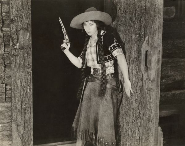 Edith Storey, in character as the female gunfighter Colonel Billy, poses with her Remington revolver in the open door of a log building in a publicity still for "As the Sun Went Down." She wears full western costume including a Mexican vest, wide belt, and a hat with a huge brim.
