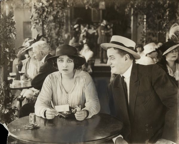 Pauline Frederick and an unidentified actor in a straw boater hat sit at a table in a cabaret in a scene still for "Ashes of Embers." She holds a wad of money in one hand and a piece of paper in the other.
