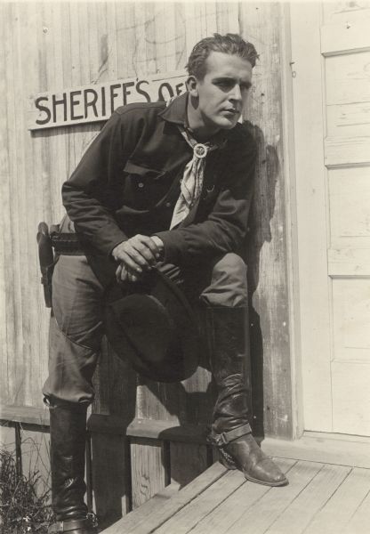 In this publicity still for the silent western "At Cripple Creek," Wallace Reid is in costume for the role of Joe Mayfield. He wears a bandanna for a tie, has a pistol on his hip, a dark wide-brimmed hat in his hand, and a sheriff's star pinned to his shirt.