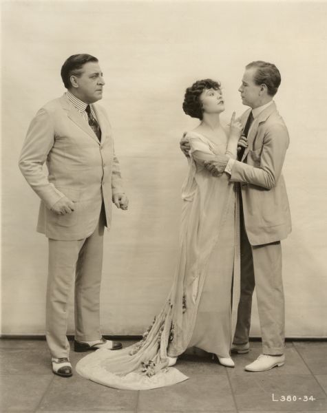 The banker William Blane (played by Joseph Kilgour) is standing with clenched fists watching his wife Cherry O'Day (Betty Compson) in the embrace of Harvey Gates (Casson Ferguson) in a publicity still for the silent drama "At the End of the World." All are dressed for summer, the men in light-colored suits, Compson in a flowing gown with a train.