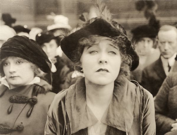 Ethel Clayton, playing Ruth Wingate, is shown in a head and shoulders close-up in a scene still for "The Attorney for the Defense." The crowd seated around seem to be spectators watching a court trial.