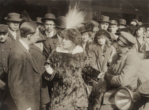 Ethel Clayton, playing Ruth Wingate, stands between a cab driver or chauffeur and another man to whom she speaks, who may well be the actor John Ince. A crowd is gathered behind them near the entrance to a public building, perhaps a train station, in this scene still for the 1915 Lubin production "The Attorney for the Defense."
