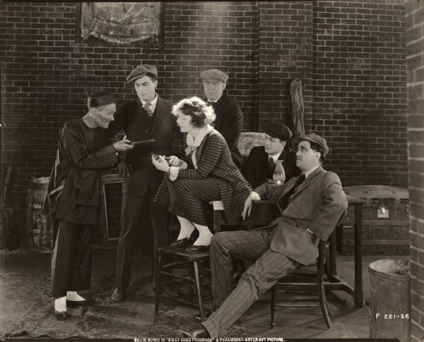 Sitting on a table in a filthy cellar, Prudence Thorne (played by Billie Burke) compares knives with a Chinaman as four other thugs watch in a scene still for "Away Goes Prudence." The Chinaman was played by M.W. Rale. The young thug standing between Rale and Burke may be played by Albert Hackett who went on to become a successful screenwriter.