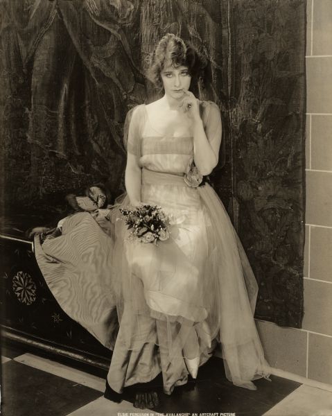 Seated on an antique chest, Elsie Ferguson models a light-colored gown, and beside her is a fur-trimmed watered-silk wrap. The French fashion house Callot Soeurs provided the costume for this publicity still for "The Avalanche."