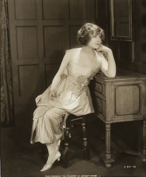 Seated on a stool with an elbow resting on a dresser, Elsie Ferguson models a white silk gown in a publicity still for "The Avalanche." The French fashion house Callot Soeurs provided the costume.