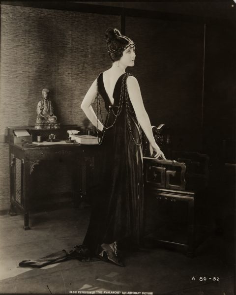 Standing looking back over her shoulder, Chichita (played by Elsie Ferguson) models a floor length black silk gown decorated with strings of black beads in a publicity still for "The Avalanche." Behind her are a chair and desk of Chinese design with a small statue of the Buddha on the desk. The French fashion house Callot Soeurs provided the costume.