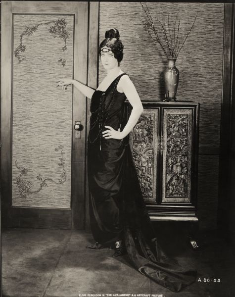 Posed by a door with her hand on her hip, Chichita (played by Elsie Ferguson) models a floor length black silk gown decorated with strings of black beads in a publicity still for "The Avalanche." Behind her is a vase on a Chinese cabinet. The French fashion house Callot Soeurs provided the costume.