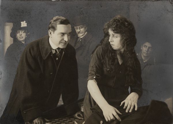 Jo, a girl of the slums played by Anita Stewart, looks over her shoulder at Roscoe Thane (Earle Williams) while, in the background, three other actors watch in a scene still for "The Awakening" (Vitagraph 1915).