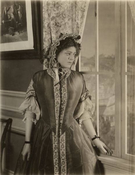 Ethel Barrymore, wearing a mid-19th century dress with basque waist and crinoline, stands by a window. She is costumed for the title role in the 1916 silent film "The Awakening of Helena Richie." On the reverse of the print, a caption in pencil reads: "Helena decides to go to the village in search of Pryor's letter."