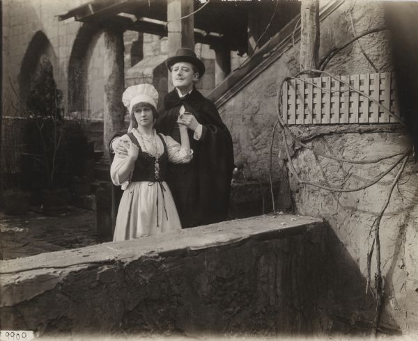 Standing in a courtyard, Raveau (played by Marc MacDermott), wearing a black cape and homburg hat, embraces Babette (Peggy Hyland) who wears the costume of a French country girl with a long skirt, vest bodice, and tall white bonnet in this scene still for "Babette" (Vitagraph 1917).