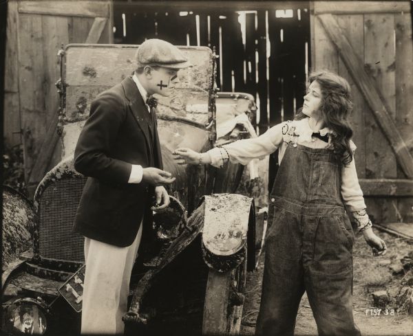 Tommy Gray (played by Richard Barthelmess) has his ring returned by Bab Archibald (Marguerite Clark) in a scene still for "Bab's Burglar." Barthelmess wears a coat, bowtie, and cloth cap and has sticking plaster or war paint on his face. Clark wears overalls. They stand in front of a muddy automobile that appears to be an Overland.