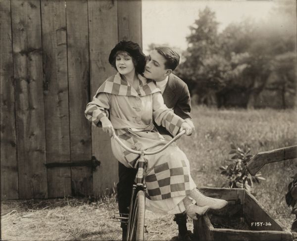 As Bab Archibald (played by Marguerite Clark) and Tommy Gray (Richard Barthelmess) 
share a bicycle, Tommy tries to steal a kiss in a scene still for "Bab's Burglar." Clark wears a boldly checked dress, large velvet beret, and white high lace-up boots.