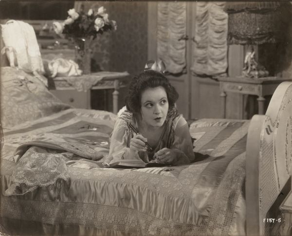 Marguerite Clark in the role of Bab Archibald lies on her bed in a nightgown, perhaps writing in a diary. This is a scene still for the 1917 Famous Players production "Bab's Burglar."