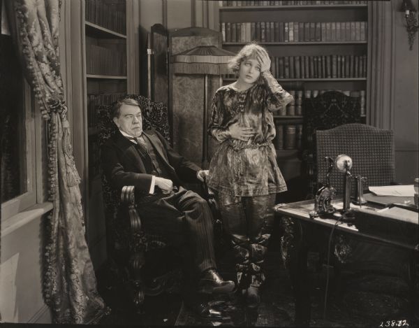 In his office at home, Senator Merrill Treadwill Marvin (played by George Fawcett) scowls suspiciously as his daughter Barbara  (Corinne Griffith) feigns sickness in a scene still for "Bab's Candidate." Griffith wears metallic satin pajamas.