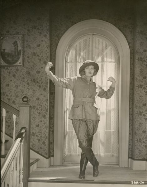 Barbara Marvin (played by Corinne Griffith) stands on a landing and flexes her biceps in a scene still for "Bab's Candidate" (Vitagraph 1920). She wears baggy trousers, a safari jacket with patch pockets, a floppy hat, and wading boots.