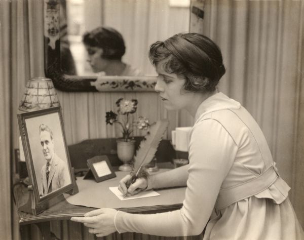 In this scene still for the 1917 Goldwyn production "Baby Mine," Zoie (played by Madge Kennedy) sits at a writing desk and gazes at a photograph of her husband Alfred (Frank Morgan).