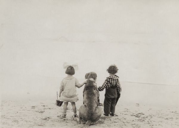With their backs to the camera, Olive Johnson, Teddy the Dog, and Francis Carpenter stand on the shore and look out to sea in this scene still for the 1915 Reliance silent short "The Baby."