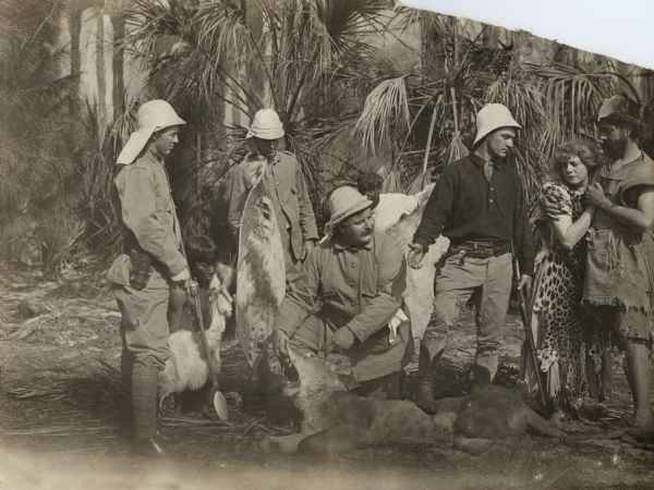 In this scene still from the 1911 Selig silent film "Back to the Primitive," a group of four men dressed for safari (pith helmets, jungle jackets, puttees) aided by two natives (who appear to be played by American Indians) have killed a lion in order to rescue a man and a woman dressed as castaways in ragged clothes and animal skins. The young man in pith helmet and dark shirt is John Wilton, played by Tom Mix. His sister, Helen Wilton, played by Kathlyn Williams, wears a leopard skin and is embraced by Will Carson, played by Charles Clary, in ragged clothes.