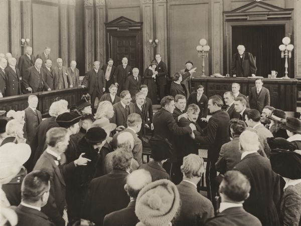 Every person in the crowded courtroom is standing in this scene still for the silent drama "Back of the Man." The center of attention is a man with a moustache, very probably the murderous Sid Wilson, played by Jack Livingston, who is being subdued by several men. In the center background a young couple embraces. They are the characters Ellen Holton and Larry Thomas, played by Dorothy Dalton and Charles Ray.