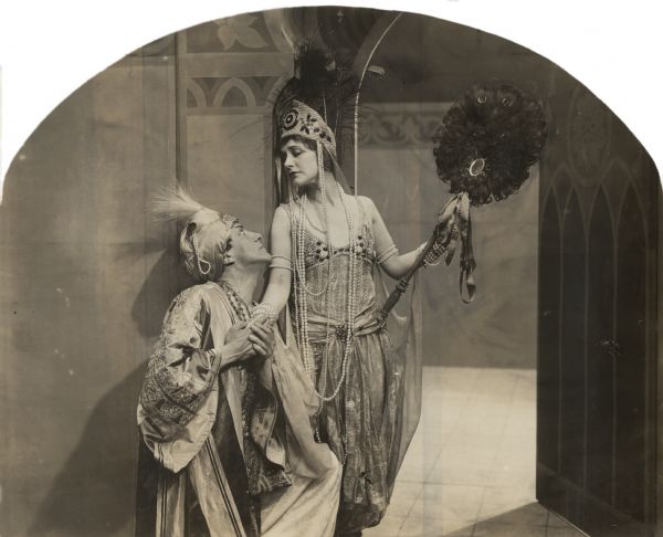 The Arab chieftan Benchaalal (played by Pedro de Cordoba) romances the English noblewoman Lady Wyverne (Elsie Ferguson) in a scene still for the silent film "Barbary Sheep." Both are costumed in Levantine splendor. Benchaalal wears a turban and silken robes. Lady Wyverne wears a complicated beaded dress, has yards of pearls around her neck, a feathered headdress on her head, and a large fan made of peacock feathers in her hand.
