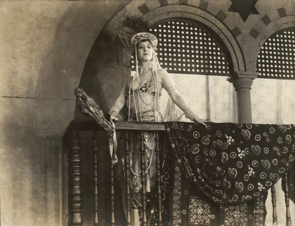 English noblewoman Lady Wyverne (played by Elsie Ferguson) poses on a balcony in a Moorish palace in a scene still for the silent film "Barbary Sheep." She wears a complicated beaded dress, has yards of pearls around her neck, a feathered headdress on her head, and a large fan made of peacock feathers in her hand.