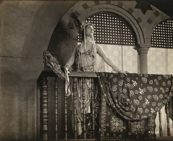 English noblewoman Lady Wyverne (played by Elsie Ferguson) poses on a balcony in a Moorish palace in a scene still for the silent film "Barbary Sheep." She wears a complicated beaded dress, has yards of pearls around her neck, a feathered headdress on her head, and a large fan made of peacock feathers in her hand. She is facing to her left.