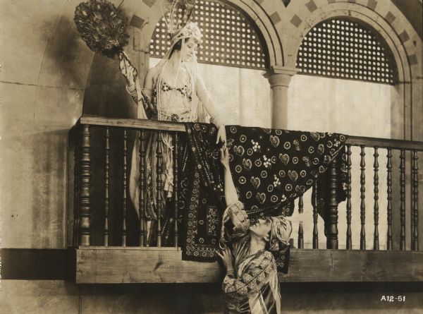 English noblewoman Lady Wyverne (played by Elsie Ferguson) poses on a balcony in a Moorish palace and offers her hand to Arab chieftan Benchaalal (Pedro de Cordoba) standing below in a scene still for the silent film "Barbary Sheep." She wears a complicated beaded dress, has yards of pearls around her neck, a feathered headdress on her head, and a large fan made of peacock feathers in her other hand. Benchaalal wears a turban and silken robes.