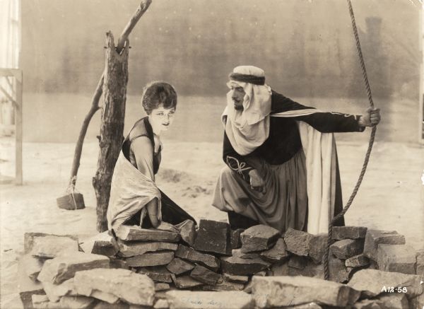 English noblewoman Kathryn, Lady Wyverne (played by Elsie Ferguson), sits on the edge of a rustic well in a scene still for the 1917 silent film "Barbary Sheep." The Arab chieftan Benchaalal (played by Pedro de Cordoba) stands with one hand holding the rope of the shaduf and looks at her intently. He wears a keffiyeh, zouave jacket and trousers, and has a Van Dyke beard.