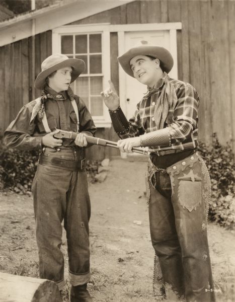 Wearing men's western clothing Jem Mason (played by Agnes Vernon) looks doubtfully at Bare Fisted Gallagher (William Desmond). She holds a Winchester repeating carbine rifle which he holds by the barrel with one hand while wagging a finger with the other. Gallagher wears charro chaps and leather wrist cuffs.