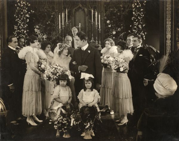 A sombre wedding party is gathered before the minister at the altar in a scene still for the silent film "The Barricade." Hope Merrill, the bride played by Mabel Taliaferro, looks seriously at her groom, John Cook, played by Clifford Bruce. The two little flower girls in the foreground look particularly unhappy.
