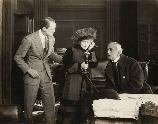 Gerald Hastings (played by Robert Rendel) listens with great interest as Hope Merrill (Mabel Taliaferro) uses a candlestick telephone by her father's desk. Her father, Amos Merrill (played by Frank Currier), is the president of a securities company. He sits at his desk chair and leans in to listen with a worried look in this scene still for the silent drama "The Barricade."